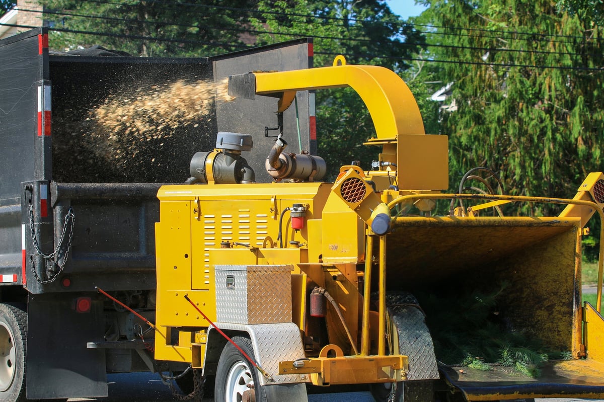 A worker stands by a machine that grinds wood while loading debris into it