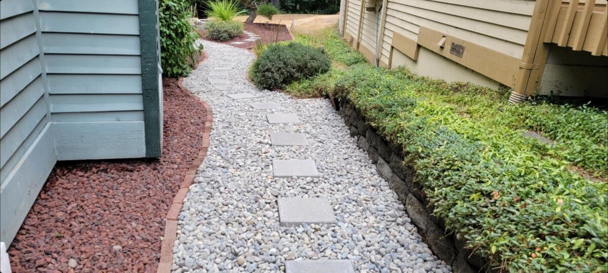 Gravel pathway between houses with red mulch on left, gray stones in center, and green hedge on right