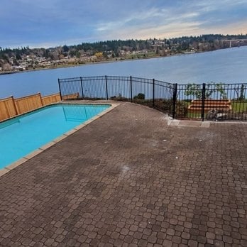 Curved swimming pool on a brick paver patio overlooking a lake with forested hills